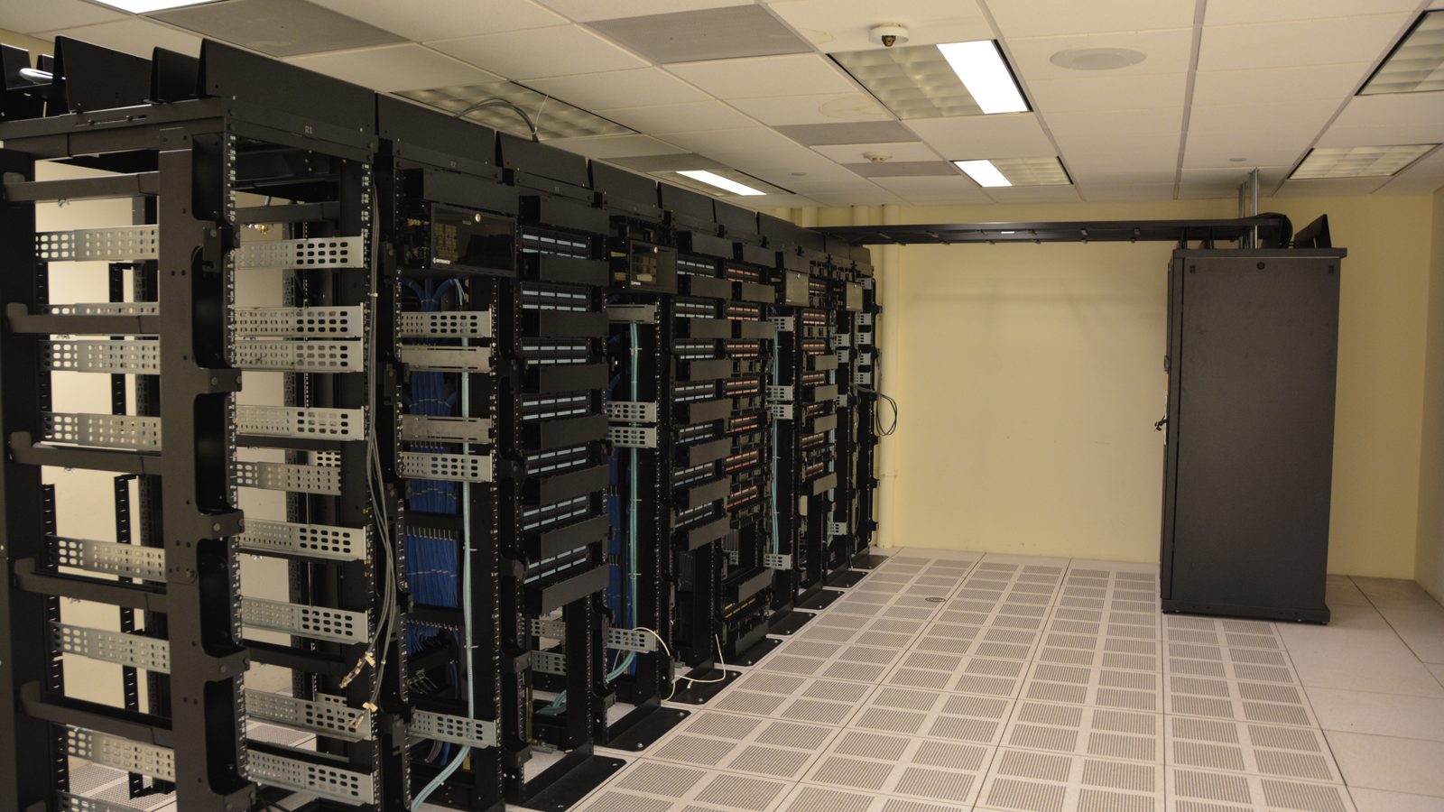 Corredor de sala de servidores com racks abertos de um lado, piso elevado perfurado e um armário de refrigeração ao fundo